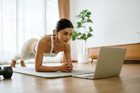 Young Asian woman in sportswear doing plank on training mat while watching online workout tutorials on laptop in the living room. Sport and recreation conceptの写真素材