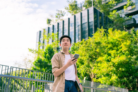Portrait of handsome Asian student using smartphone. Young man standing outdoor happy smiling with holding mobile phoneの写真素材