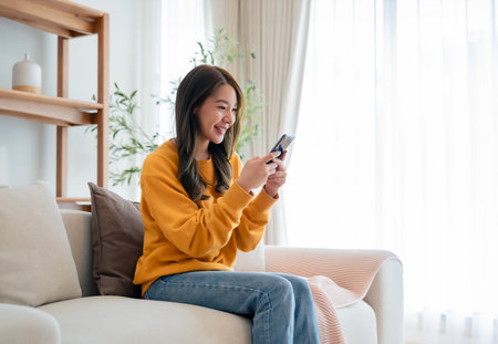 Happy young asian woman relax on comfortable couch at home texting messaging on smartphone, smiling girl use cellphone chatting, browse wireless internet on gadget, shopping online from homeの写真素材