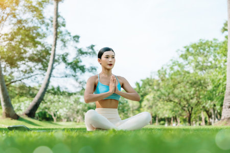 Asian woman meditating in a peaceful park setting, sitting cross-legged on green grass with trees in the background on a sunny day. Concept of mindfulness, self-care, and outdoor relaxationの写真素材