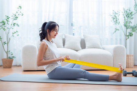 Young Asian woman doing pilates workout using elastic strap pulling with arms for shoulder training on yoga mat indoors. Resistance band exercise at homeの写真素材