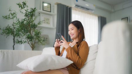 Happy young asian woman relax on comfortable couch at home texting messaging on smartphone, smiling girl use cellphone, chatting online message, shopping online from homeの写真素材
