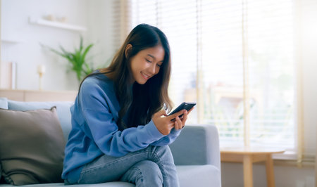Happy young asian woman relax on comfortable couch at home, typing chat message on smartphone, smiling girl use cell phone chatting, searching information on browser wireless internet, online shoppingの写真素材