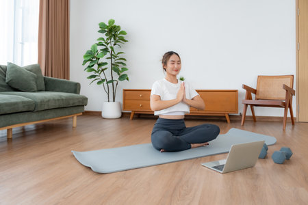 Young Asian woman meditating in lotus position at home, sitting on the floor in fitness clothing. Breathing workout to achieve relaxation, peace and mindfulnessの写真素材
