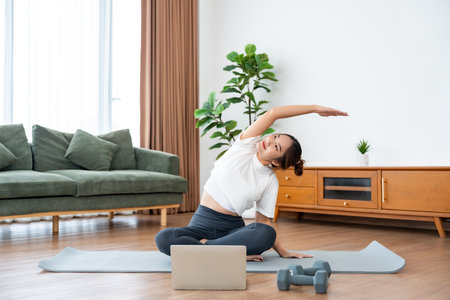 Young Asian woman stretching yoga workout on exercise mat while online training class with computer laptop in living roomの写真素材