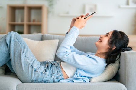 Happy cheerful positive young Asian woman on the phone at home, smiling girl using app and browsing on the internet on smartphone lying on sofaの写真素材