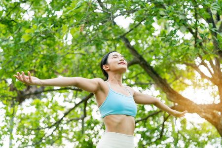 Young Asian woman relaxed breathing fresh air with trees in the background on a sunny day. Concept of wellness, healthy lifestyle, mindfulness, and outdoor fitnessの写真素材