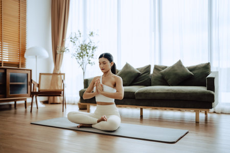 Young Asian woman meditating in lotus position at home, sitting on the floor in fitness clothing. Breathing workout to achieve relaxation, peace and mindfulnessの写真素材