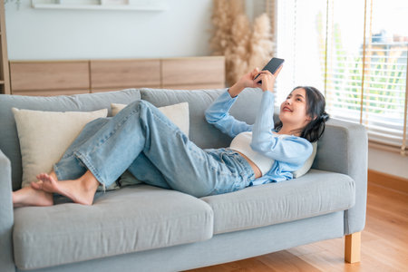 Happy cheerful positive young Asian woman on the phone at home, smiling girl using app and browsing on the internet on smartphone lying on sofaの写真素材