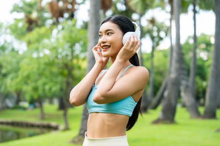 Asian sports woman listening to music on headphones outdoors. Female in sportswear standing against in park. Happy lady athlete taking a break from her workoutの写真素材