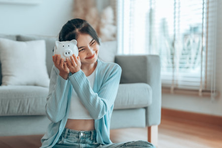 Young Asian woman holding piggy bank and sitting on the floor at home. Save money and financial investmentの写真素材