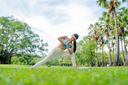 Asian woman practicing yoga outdoors in a peaceful green park, performing a twisting side lunge pose with hands in prayer position. Promoting mindfulness, flexibility, and a healthy lifestyleの写真素材