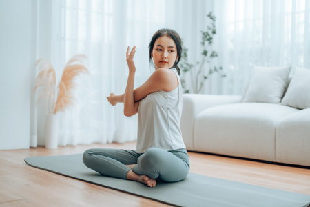 Happy young Asian woman practicing yoga and shoulder stretching at home sitting on floor in living roomの写真素材