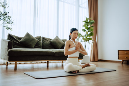 Young Asian woman meditating in lotus position at home, sitting on the floor in fitness clothing. Breathing workout to achieve relaxation, peace and mindfulnessの写真素材