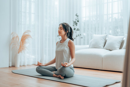 Young Asian woman meditating in lotus position at home, sitting on the floor in fitness clothing. Breathing workout to achieve relaxation, peace and mindfulnessの写真素材
