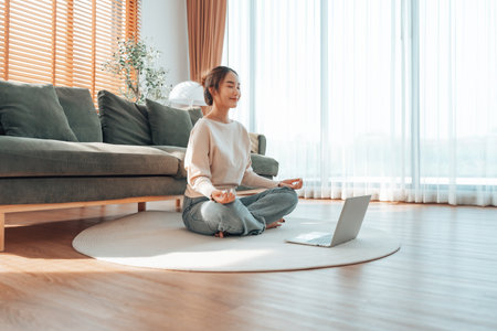 Happy young Asian woman practicing yoga and meditation at home sitting on floor in living room in lotus position with laptopの写真素材