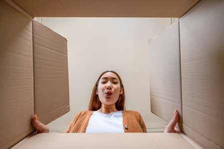 Low angle view of surprised young asian woman unpacking. Opening carton box and looking inside....