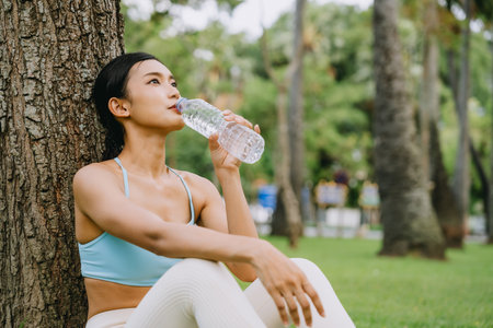 Young Asian woman in activewear sitting under a tree, drinking water after a workout in the park. She looks refreshed and relaxed, enjoying hydration and nature in a peaceful outdoor environmentの写真素材