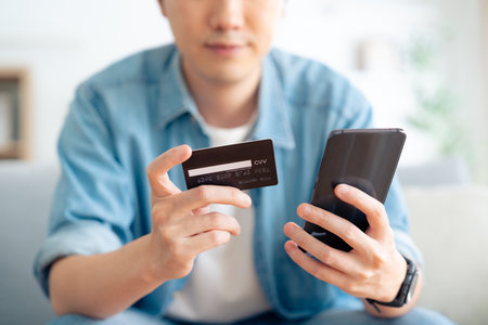 Young asian man sitting on sofa in living room, makes online banking payments through the internet from bank card on cell phone. Shopping online on mobile with credit cardの写真素材
