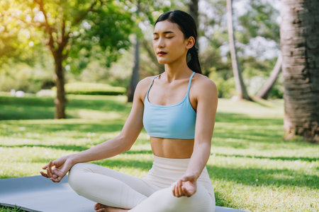 Asian woman meditating in a peaceful park setting, sitting cross-legged on green grass with trees in the background on a sunny day. Concept of mindfulness, self-care, and outdoor relaxationの写真素材