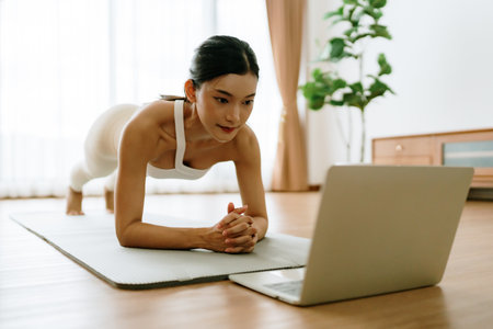 Young Asian woman in sportswear doing plank on training mat while watching online workout tutorials on laptop in the living room. Sport and recreation conceptの写真素材
