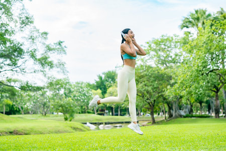 Young Asian woman in sportswear jumping happily during outdoor workout in a green park. Active lifestyle, fitness, and wellness concept in natureの写真素材
