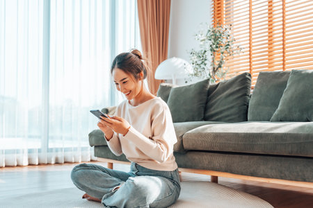 Smiling pretty young Asian woman sitting on cozy couch, using funny mobile apps in living room. Woman at Home, Doing Online Shopping, Messaging Friends, Posting on Social Mediaの写真素材