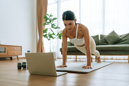 Young Asian woman in sportswear doing plank on training mat while watching online workout tutorials on laptop in the living room. Sport and recreation conceptの写真素材