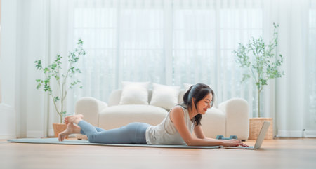 Young Asian woman lying on the floor on yoga mat in front of laptop and training at home. Beautiful smiling woman doing stretching exercise while communicates with a trainer online before workoutの写真素材