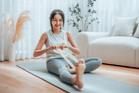 Young Asian woman doing pilates workout using elastic strap pulling with arms for shoulder training on yoga mat indoors. Resistance band exercise at homeの写真素材