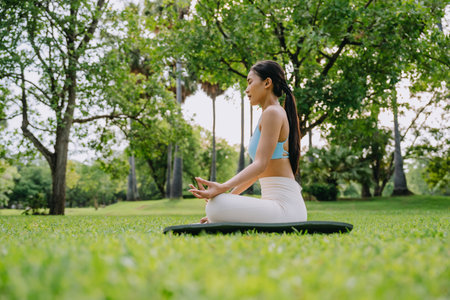 Asian woman meditating in a peaceful park setting, sitting cross-legged on green grass with trees in the background on a sunny day. Concept of mindfulness, self-care, and outdoor relaxationの写真素材