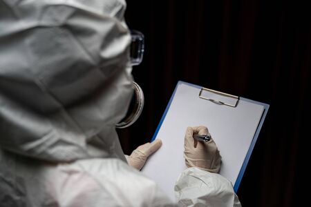 Woman wearing gloves with biohazard chemical protective suit and mask. is writing report on paper.