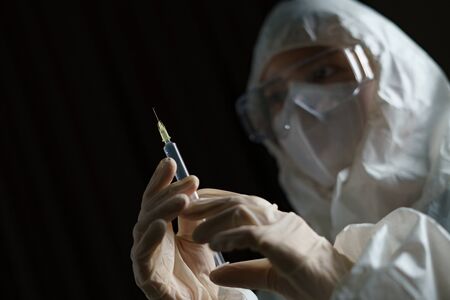 Woman wearing gloves with biohazard protective suit and mask holds an virus vaccine with syringe,...