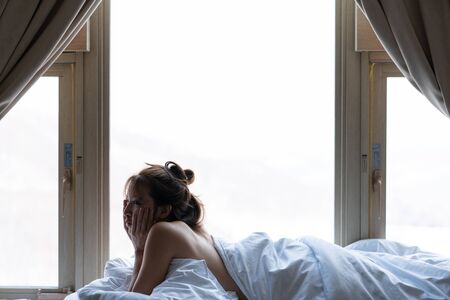 Woman sits on a window sill wrapped in a warm blanket, relaxing on cozy bed in log cabin.の写真素材