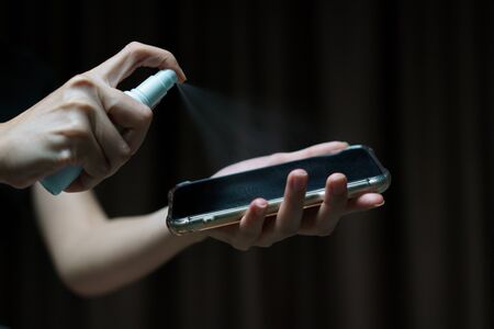 Hand holding and cleaning mobile phone screen with isopropyl alcohol spray  for corona virus or...