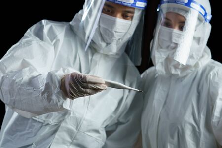 Staff wearing ppe suit and face mask and face shield, looking in thermometer. Corona virus, Covid-19 concept.の写真素材