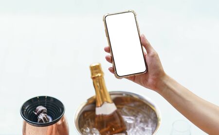 Woman hand holding blank white screen mobile phone with champagne bottle in ice bucket and two glasses near bubble poolの写真素材