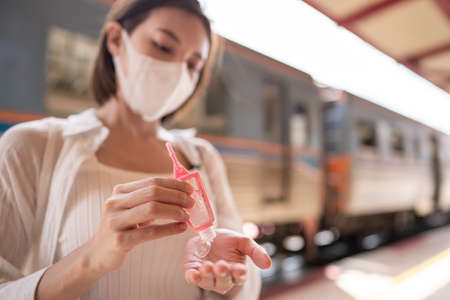 Woman in protective mask using hand sanitizer gel during wait for the local train on platform at station.の写真素材