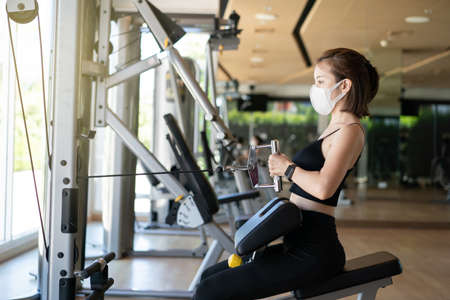 Woman wearing face mask seated cable row, pulling cable of rowing machine training in gym. during...