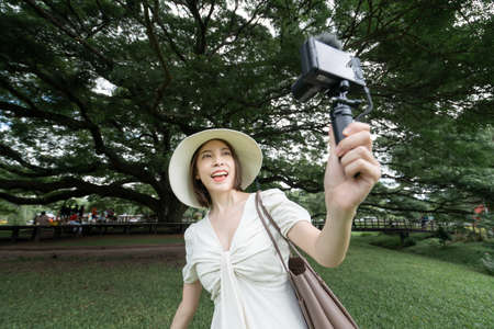 Woman wearing white dress taking selfie under the Giant Monkey Pod trees in Kanchanaburi, Thailand.