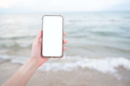 Woman hand holding mobile phone with blank white screen mockup on the beach.の写真素材