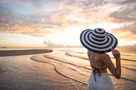 Woman in top bikini and white long pant wearing hat on the beach with a beautiful sunrise or sunset in background.の写真素材