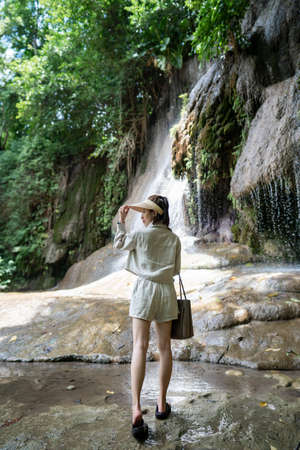 Back side of woman with waterfall in tropical rainforest with rock. Saiyok noi Waterfall, Located in Kanchanaburi province, Thailand.の写真素材
