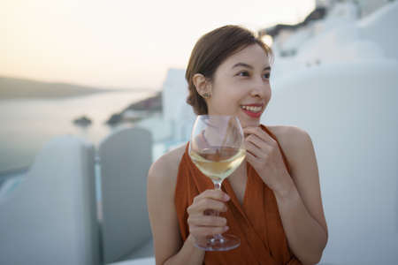 Woman tourist in gorgeous dress with glass of wine relaxing at sunset view at whitewashed village in Oia, Santorini Greece. mediterranean sea.の写真素材