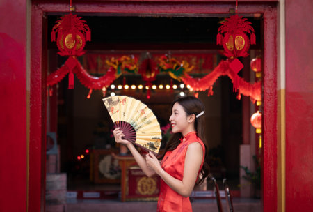 Asian woman wearing red Cheongsam dress and holding a golden fan at Chinese shrine. Chinese new year...