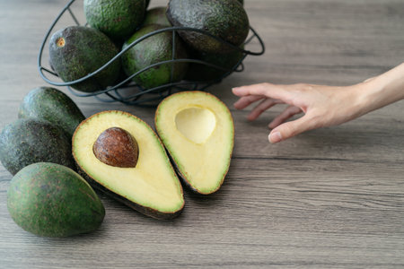 Woman hand with cut half, sliced fresh green avocado on brown wooden table. Fruits healthy food...