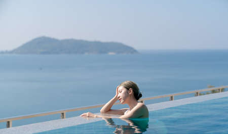 Woman in white bikini relaxing at the edge of an infinity pool with the view over the blue sea.