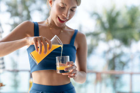 Asian woman pouring orange juice in to the glass, pool and garden background.