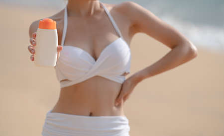 Woman in white bikini holding sunscreen bottle in hand on the beach.の写真素材