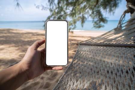 Hand holding mock up mobile with white sceen while laying on hammock swing between trees on the beach.の写真素材